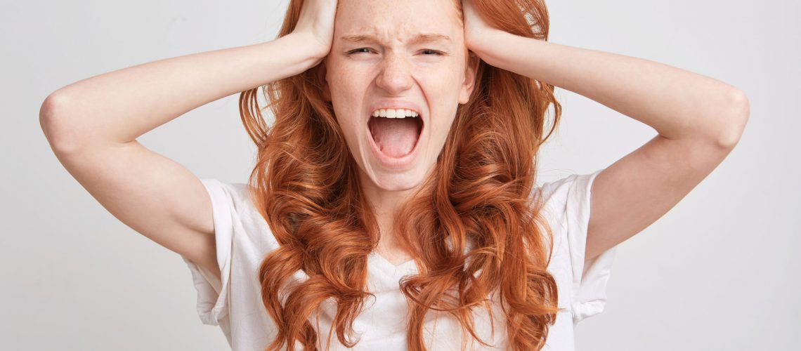 Portrait of desperate hysterical young woman with red wavy long hair, freckles and opened mouth wears t shirt feels angry and shouting isolated over white background